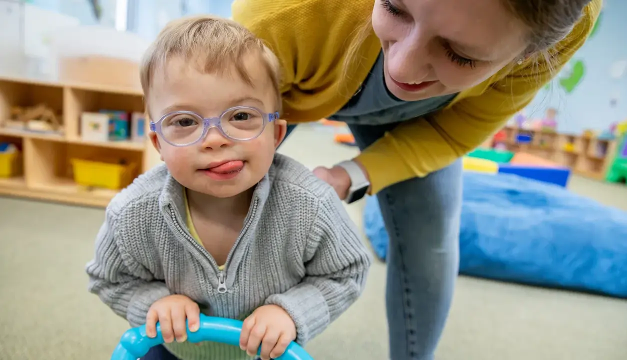 Chłopiec z zespołem Downa, w okularach, z językiem na brodzie, bawi się z terapeutką. To przykład pracy w obszarze oligofrenopedagogiki.