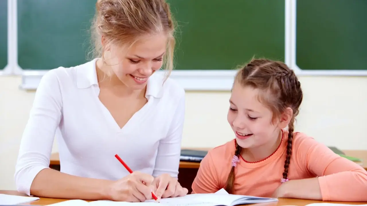 Uśmiechnięta nauczycielka wspomagająca z kwalifikacjami pomaga uczennicy w lekcjach przy biurku.