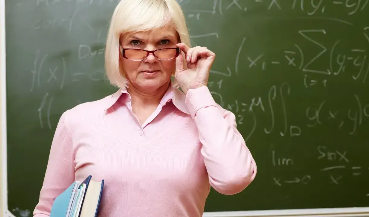 Nauczycielka matematyki poprawia okulary, trzymając książki. Na tablicy widać wzory. Dziś jest 14 października.