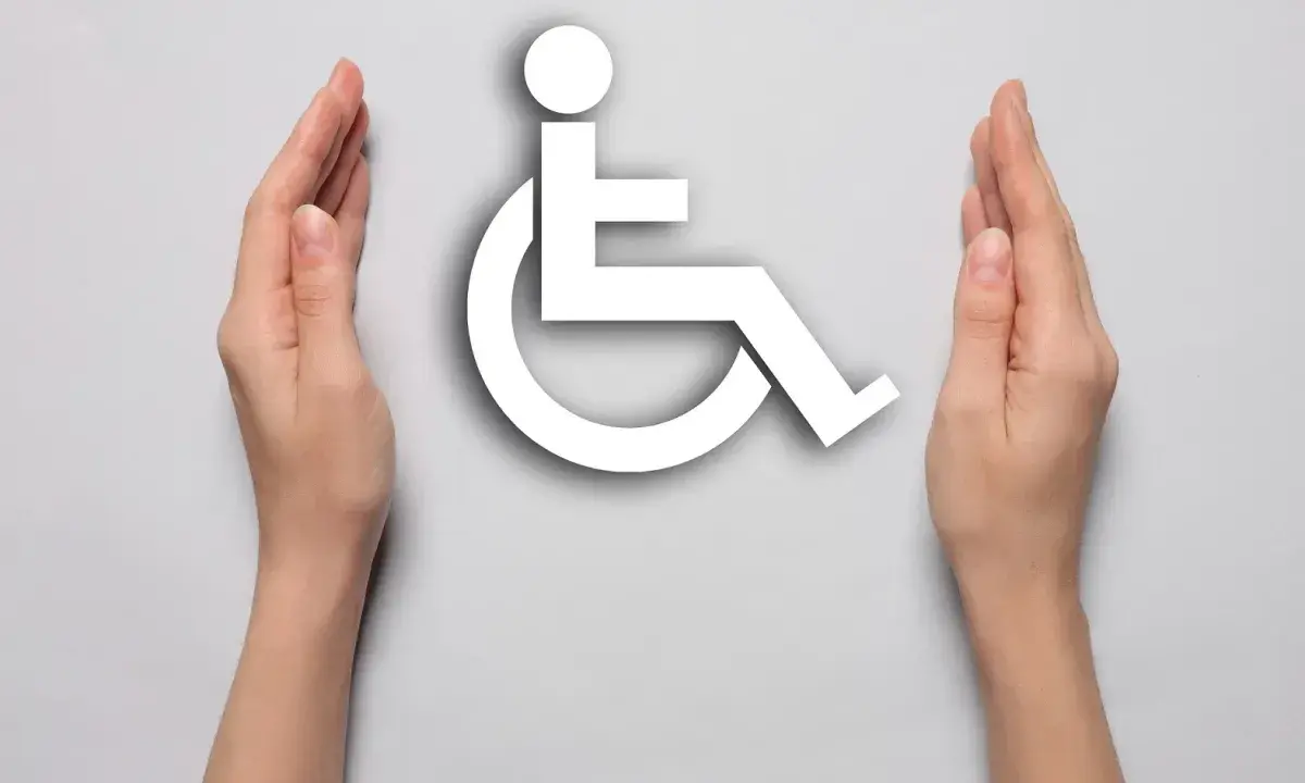 Dwie dłonie otaczają biały symbol osoby na wózku inwalidzkim, symbolizując wsparcie i akceptację dla osób z niepełnosprawnościami.