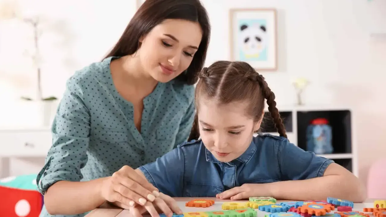Kobieta pomaga dziewczynce z niepełnosprawnością intelektualną w stopniu lekkim układać kolorowe puzzle.