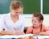 Uśmiechnięta nauczycielka wspomagająca z kwalifikacjami pomaga uczennicy w lekcjach przy biurku.