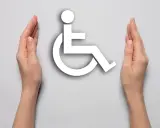Dwie dłonie otaczają biały symbol osoby na wózku inwalidzkim, symbolizując wsparcie i akceptację dla osób z niepełnosprawnościami.