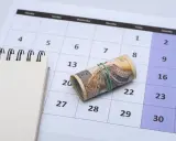 Zrolowane banknoty na kalendarzu, symbolizujące ekwiwalent czasu i pieniędzy. Obok pusty notatnik.