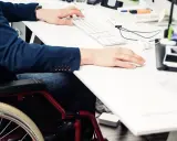 Mężczyzna w garniturze pracuje przy biurku na wózku inwalidzkim, co świadczy o jego lekki stopień niepełnosprawności.