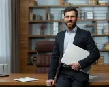 Uśmiechnięty dyrektor szkoły w garniturze, trzymający laptopa, stoi przy biurku w gabinecie z regałami pełnymi książek.