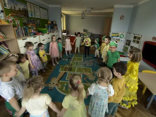 Dzieci trzymają się za ręce, tworząc krąg na dywanie z zabawnymi zwierzątkami. To radosna zabawa w **zespole szkolno przedszkolnym**.