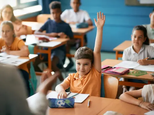 Chłopiec z podniesioną ręką czeka na ocenianie odpowiedzi. Inne dzieci pracują przy ławkach.