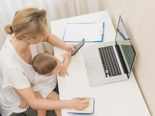 Kobieta z dzieckiem na kolanach sprawdza na telefonie, urlop macierzyński ile płatny. Obok laptop i notatnik.