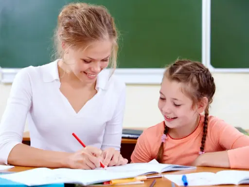 Uśmiechnięta nauczycielka wspomagająca z kwalifikacjami pomaga uczennicy w lekcjach przy biurku.