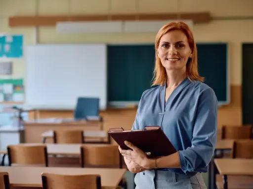 Uśmiechnięta nauczycielka z rudymi włosami trzyma teczkę. W tle klasa z ławkami i tablicą. Posiada legitymację nauczyciela.