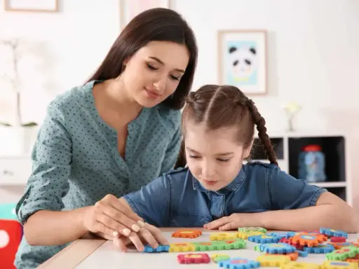 Kobieta pomaga dziewczynce z niepełnosprawnością intelektualną w stopniu lekkim układać kolorowe puzzle.