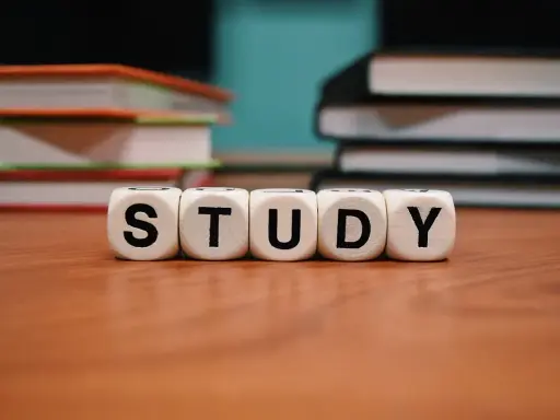 Białe kostki z literami "STUDY" na drewnianym stole, w tle stosy książek.