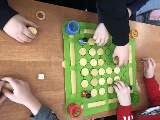 Dzieci grają w grę planszową podczas zajęć korekcyjno kompensacyjnych. Na zielonej macie są drewniane pionki i kostka.