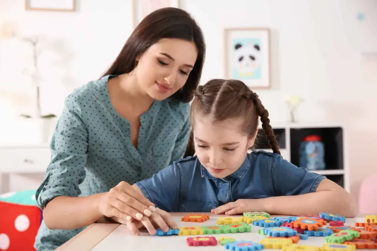 Kobieta pomaga dziewczynce z niepełnosprawnością intelektualną w stopniu lekkim układać kolorowe puzzle.