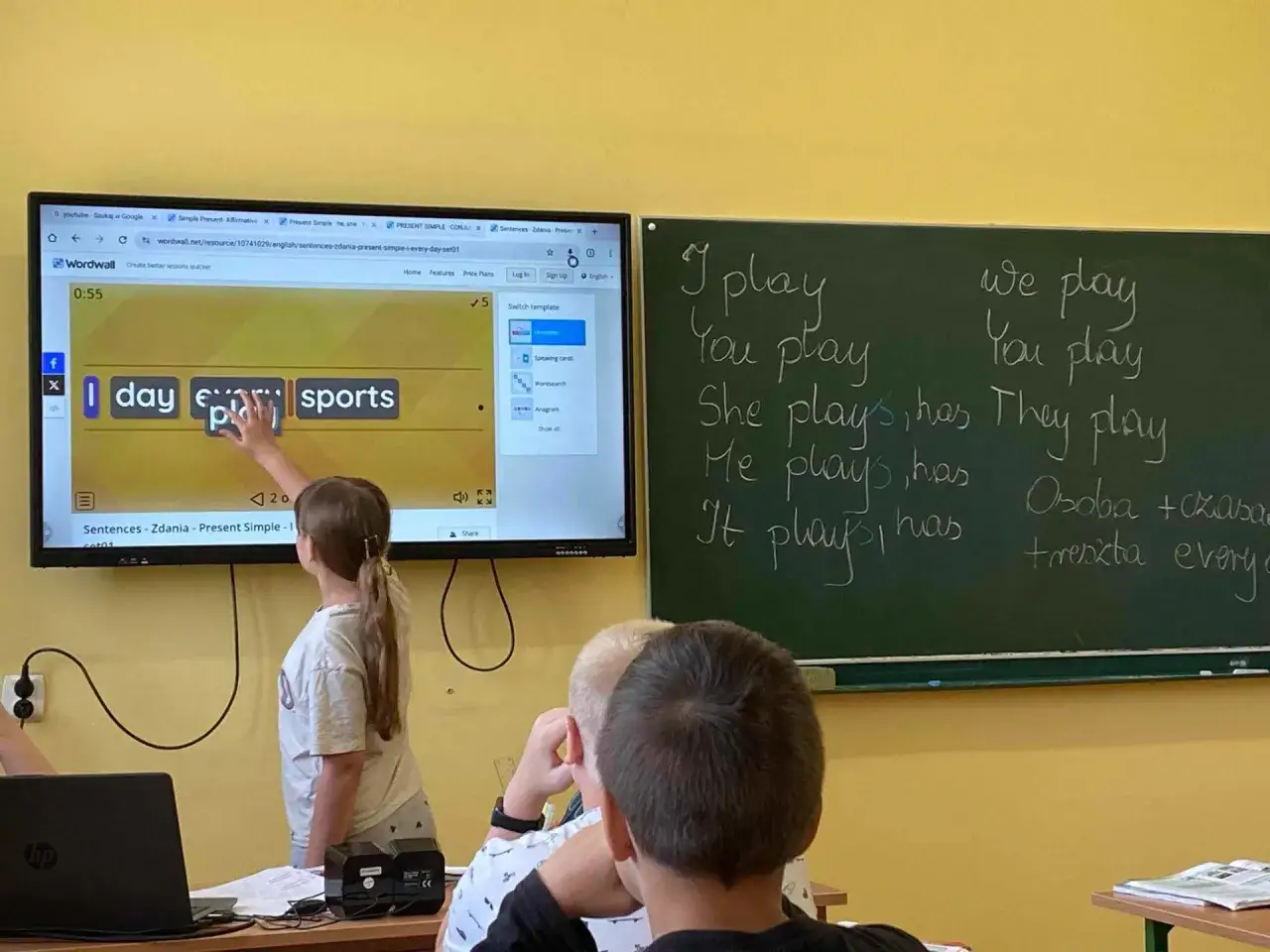 Dziewczynka interaktywną tablicą ćwiczy angielskie zdania. Na tablicy wyświetla się ćwiczenie z Wordwall, a obok zapisane są przykłady zdań.