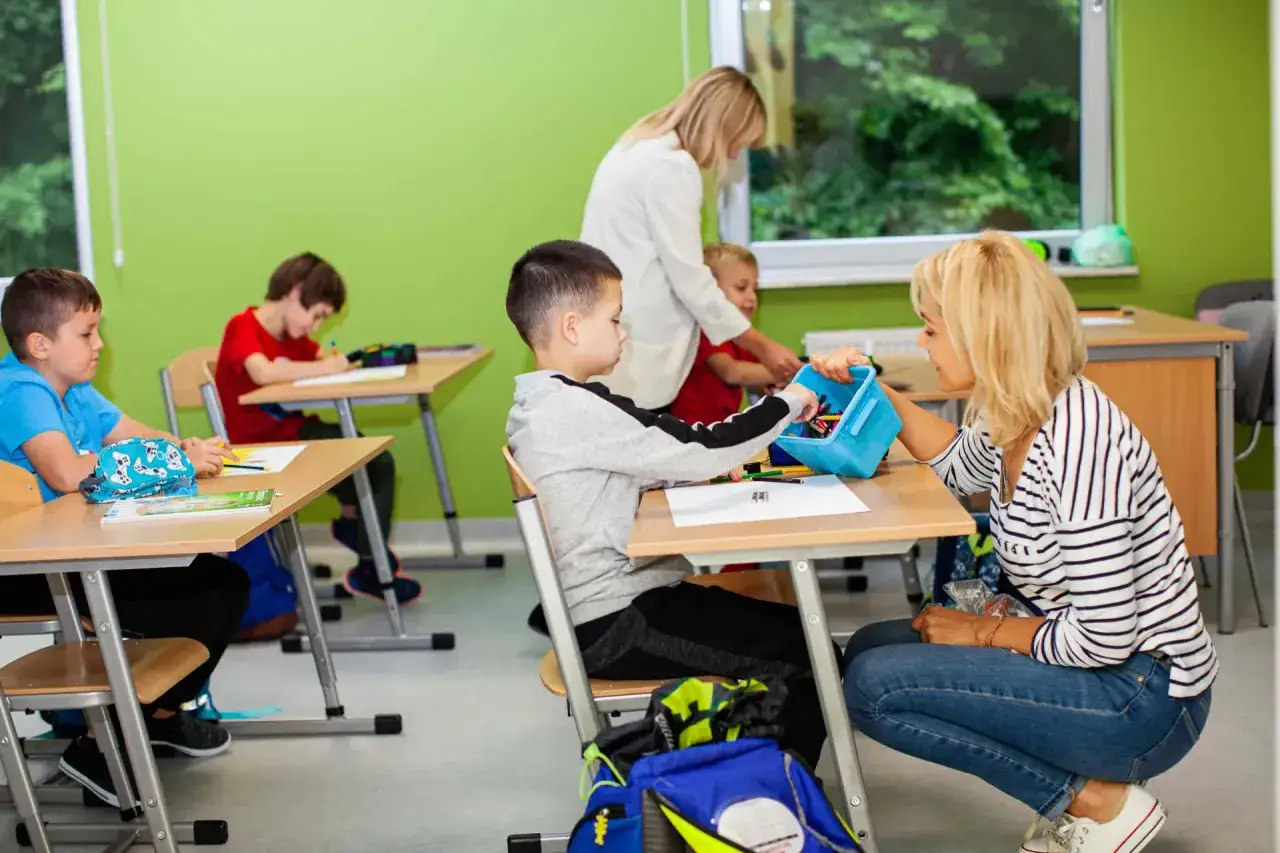 Nauczycielka pomaga chłopcu, który jest neurotypowy, wybrać kredki z niebieskiego pudełka. Dzieci siedzą przy ławkach w jasnej sali lekcyjnej.